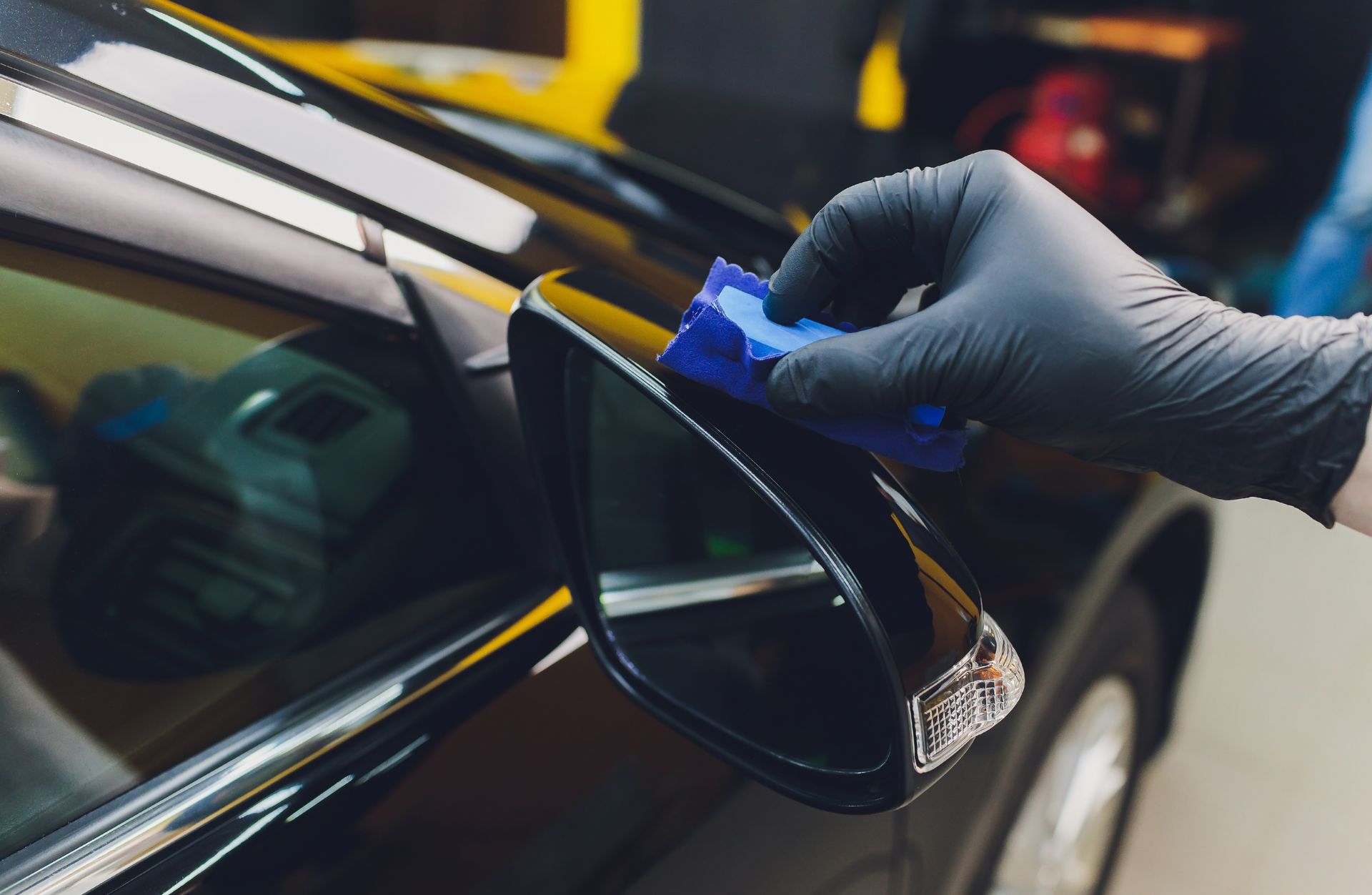Gloved hand applying blue tape to a black car’s side mirror while detailing the vehicle