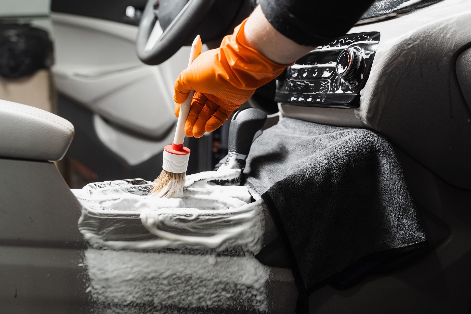 Orange-gloved hand cleaning a car seat with a brush and soap foam inside a vehicle.
