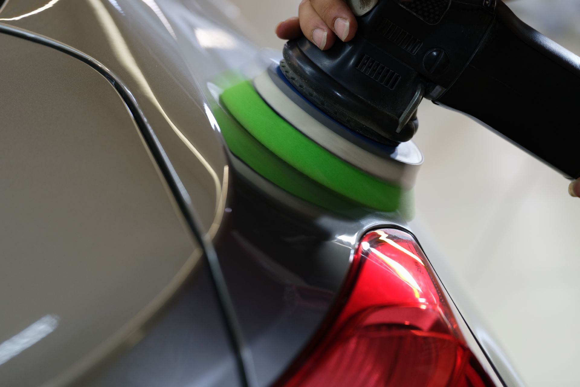 Hands buffing a black car with a green polishing pad near the red tail light