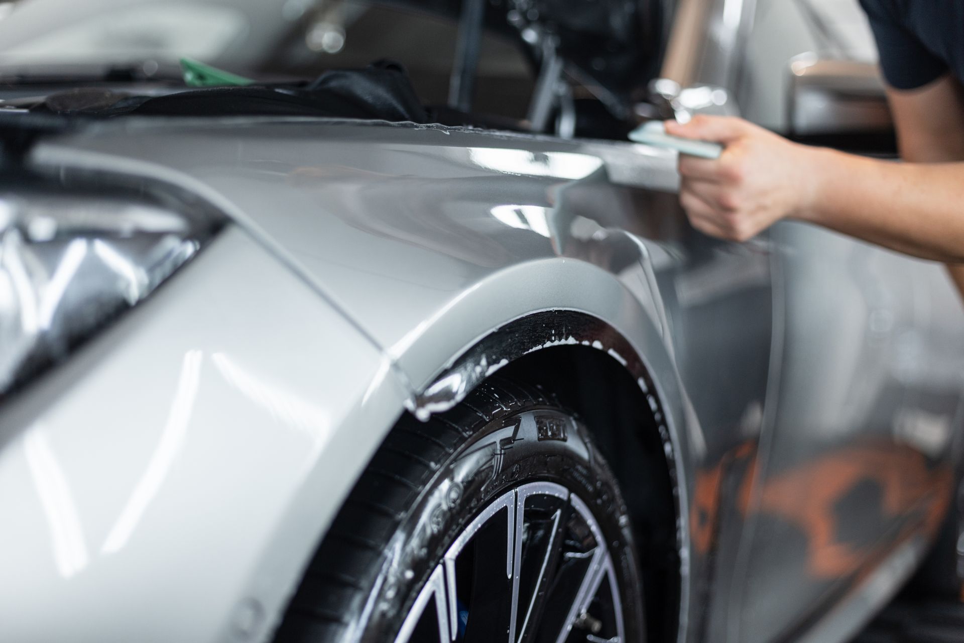 Person polishing a silver car’s fender and wheel with a cloth in a garage