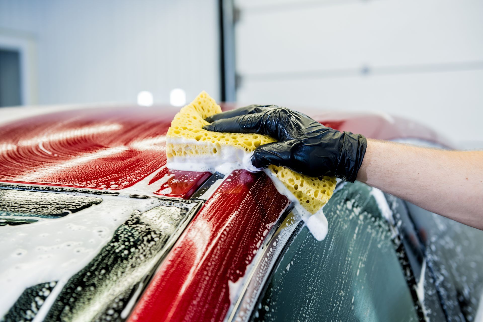 Gloved hands scrubbing soap off a red car windshield with a sponge in a car wash