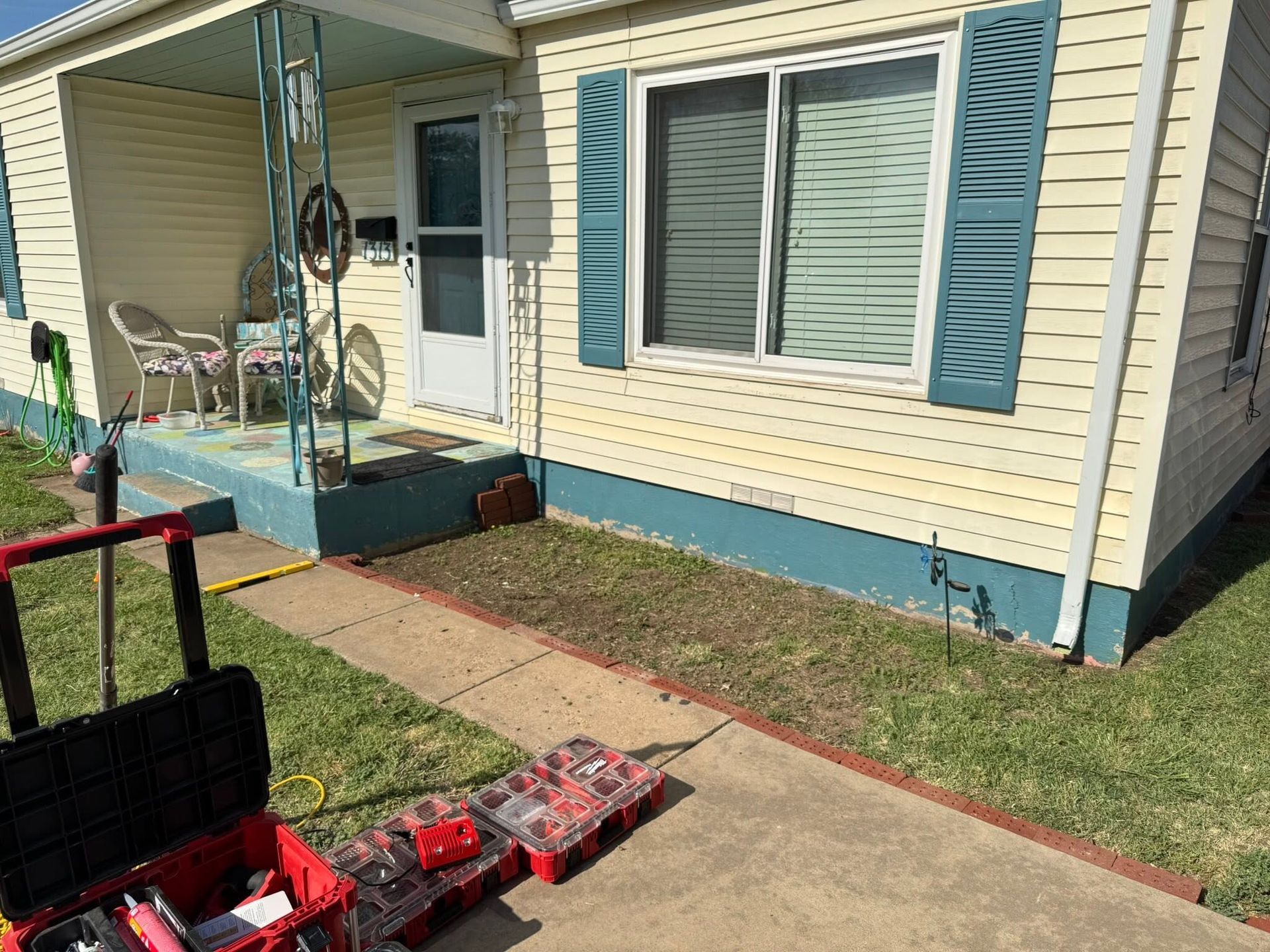 Front porch of a beige house with blue shutters, sidewalk, and a tool cart in the foreground.