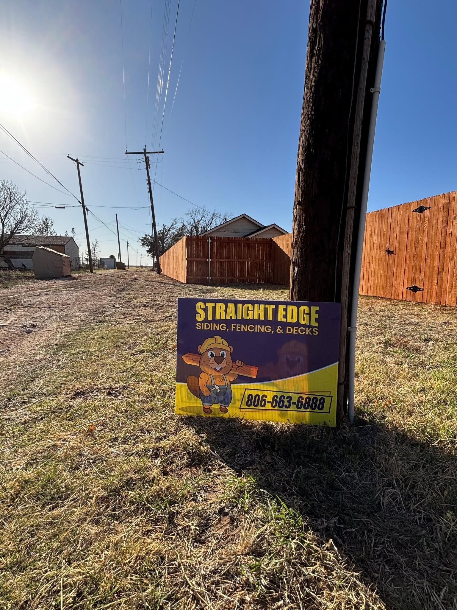Purple sign for “Stray Cat Edge” in a grassy yard beside a wooden fence under a bright sky.