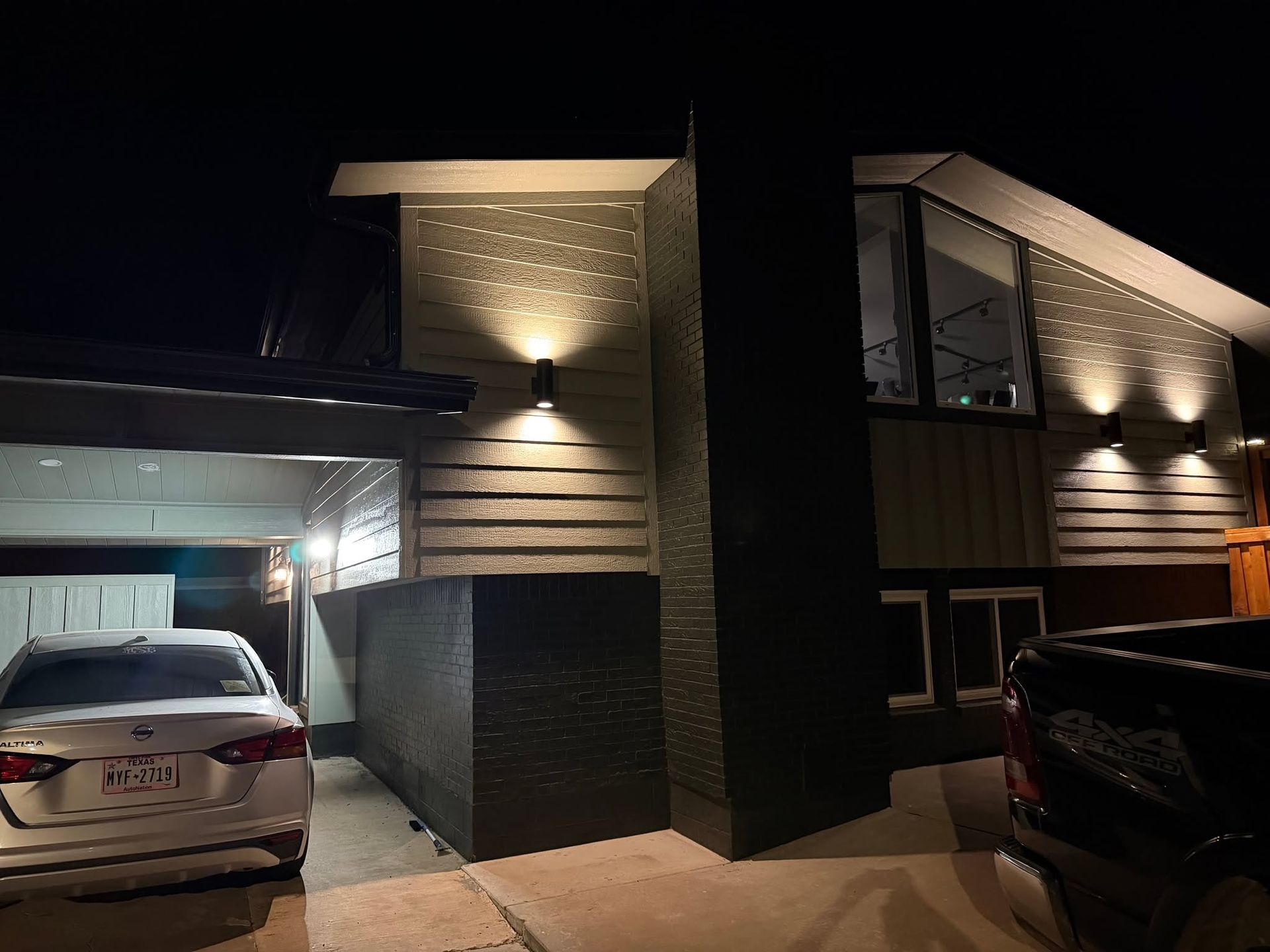 Nighttime view of a modern house exterior with lit driveway, carport, and parked cars