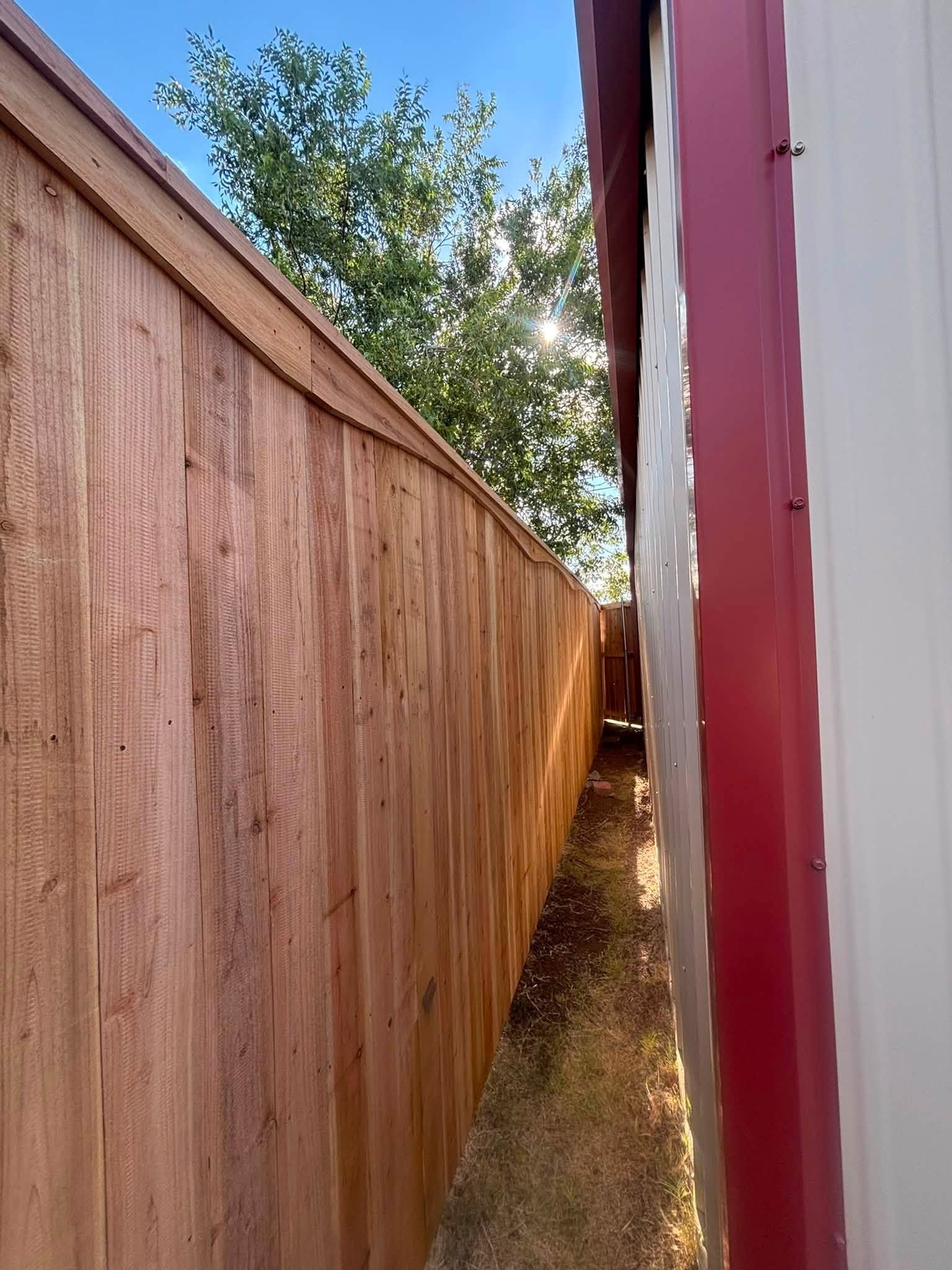 Narrow alley between a wooden fence and a red building, with sunlight and trees above.