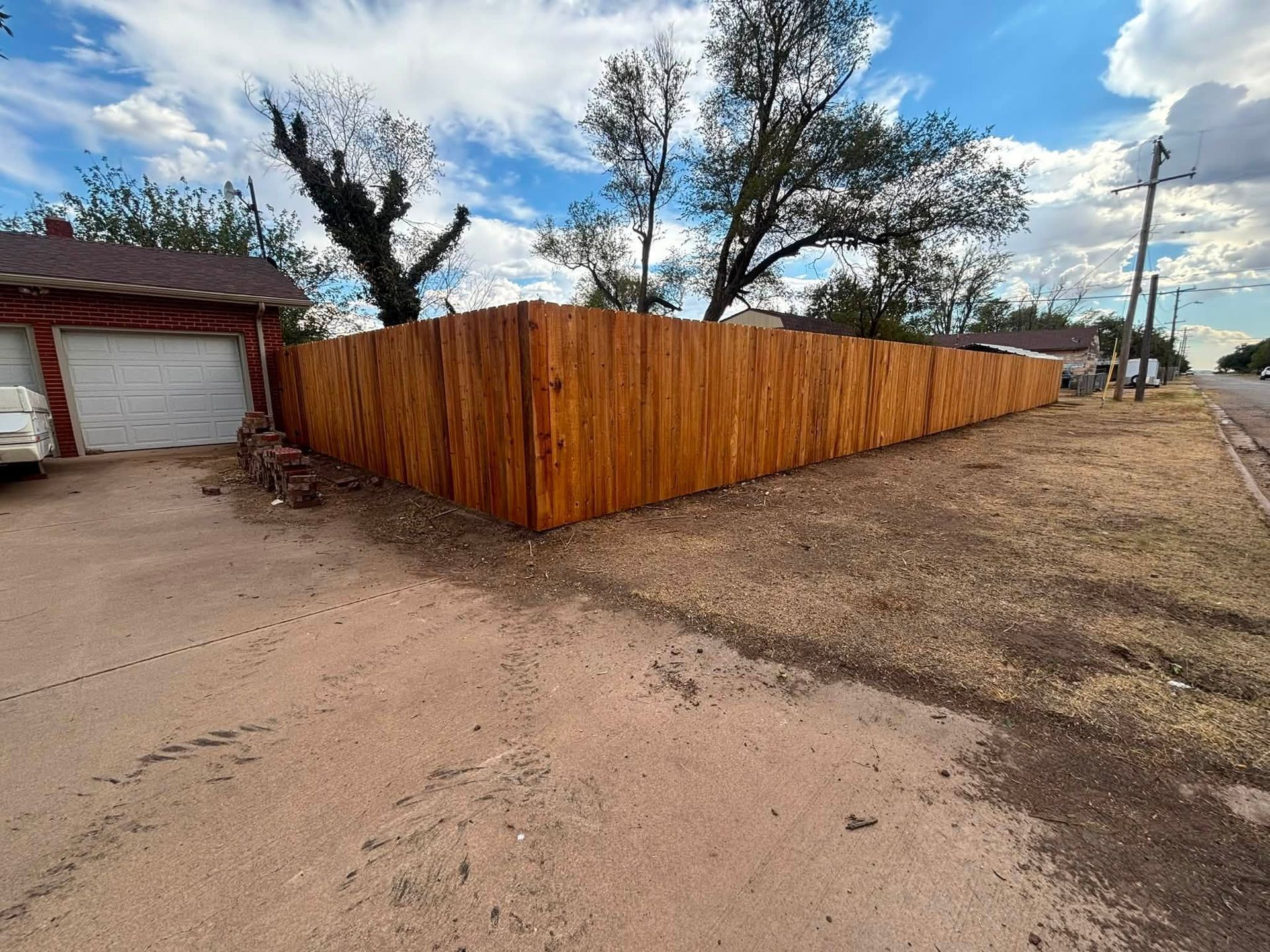 Wooden privacy fence beside a garage along a dirt roadside under a cloudy sky