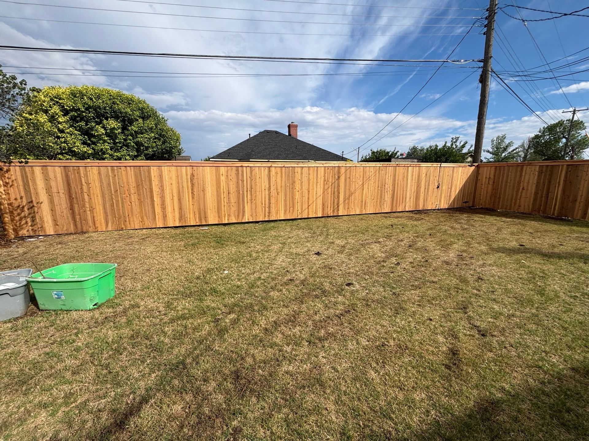 Backyard with dry grass, wooden fence, blue sky, and green and gray containers in the corner
