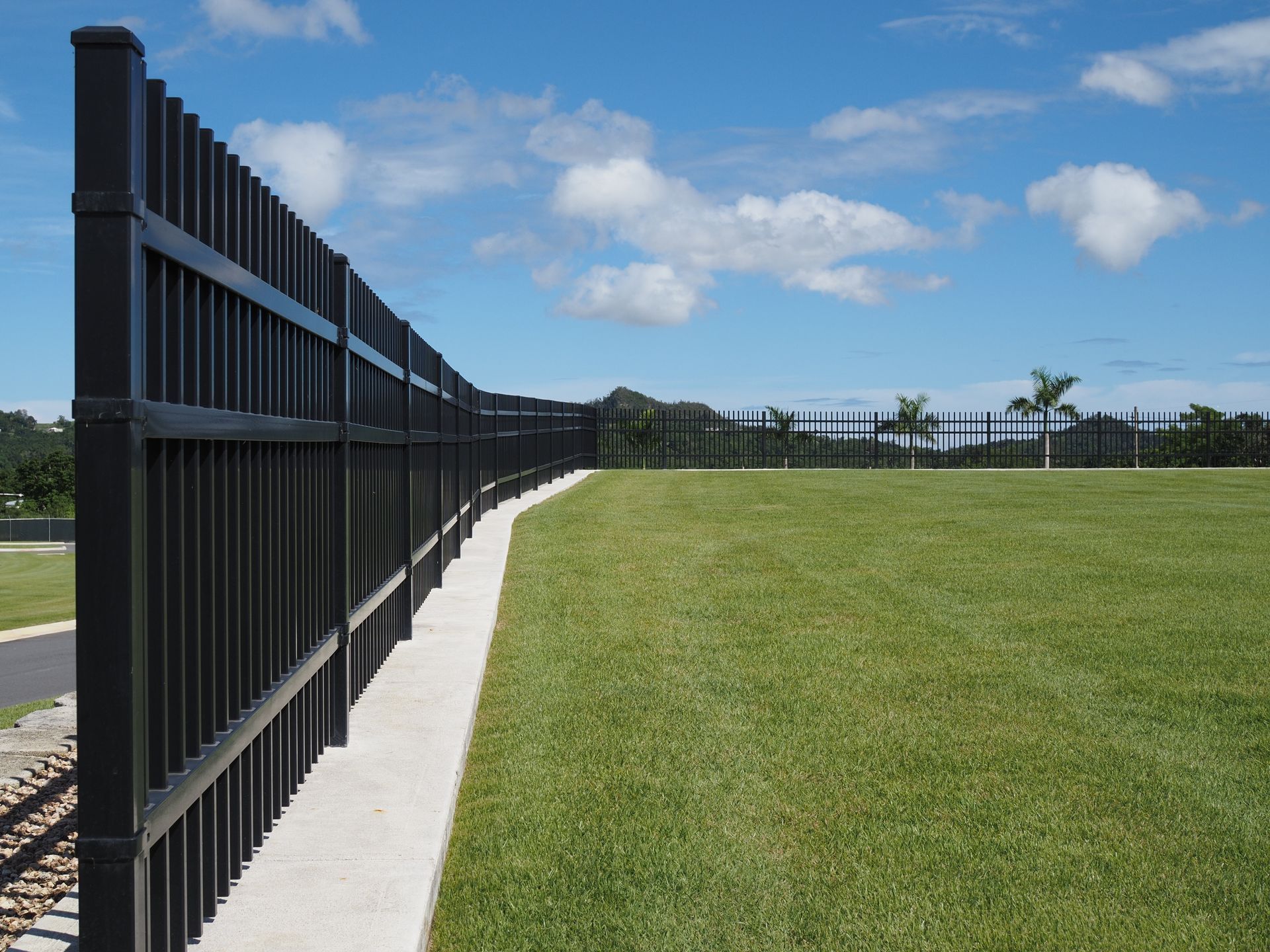 Black metal fence lines a curved concrete path beside a green lawn under a blue sky with clouds.