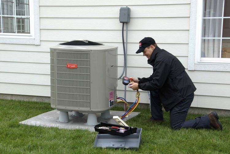 Technician kneeling beside outdoor AC unit, checking refrigerant with gauges outside a house.