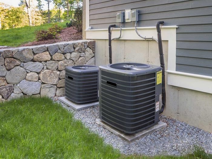 Two outdoor HVAC units beside a house, on gravel next to a stone retaining wall and green lawn