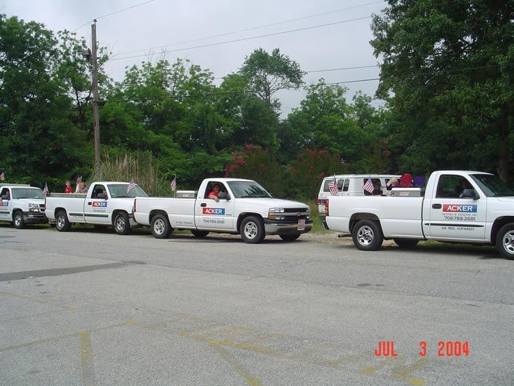 Line of white pickup trucks parked along a roadside with trees and a cloudy sky in the background.