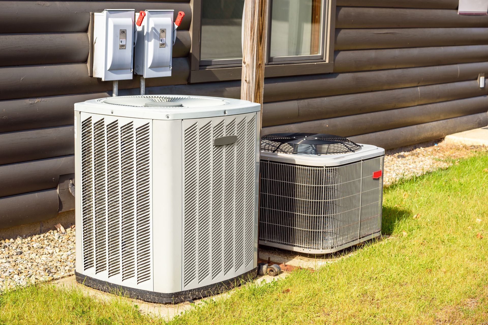 Outdoor HVAC units beside a log cabin, one white and one dark gray, on a grassy lawn.