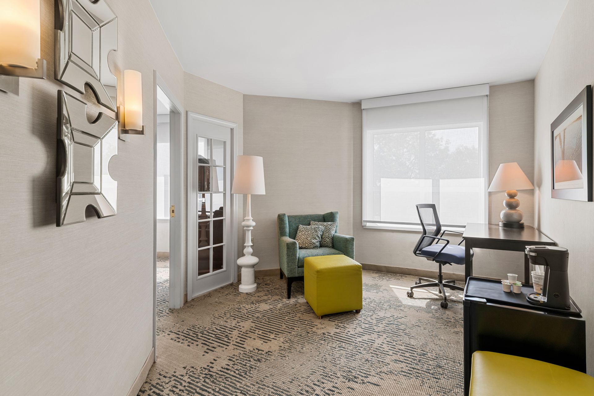 Modern home office with desk, chair, lamps, green ottoman, and patterned chair near a bright window