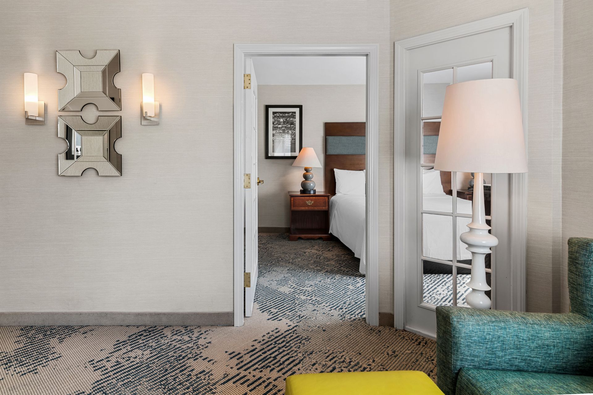 Modern hotel room view through open double doors, with patterned carpet, wall sconces, and floor lamps.