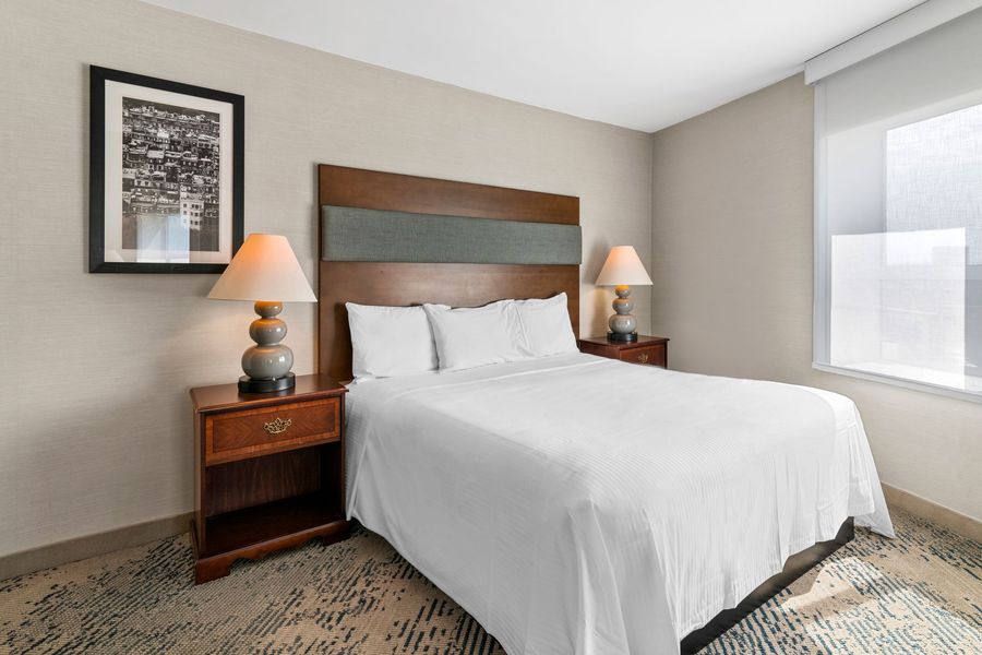 Modern hotel bedroom with white bed, wooden headboard, bedside lamps, and neutral decor.