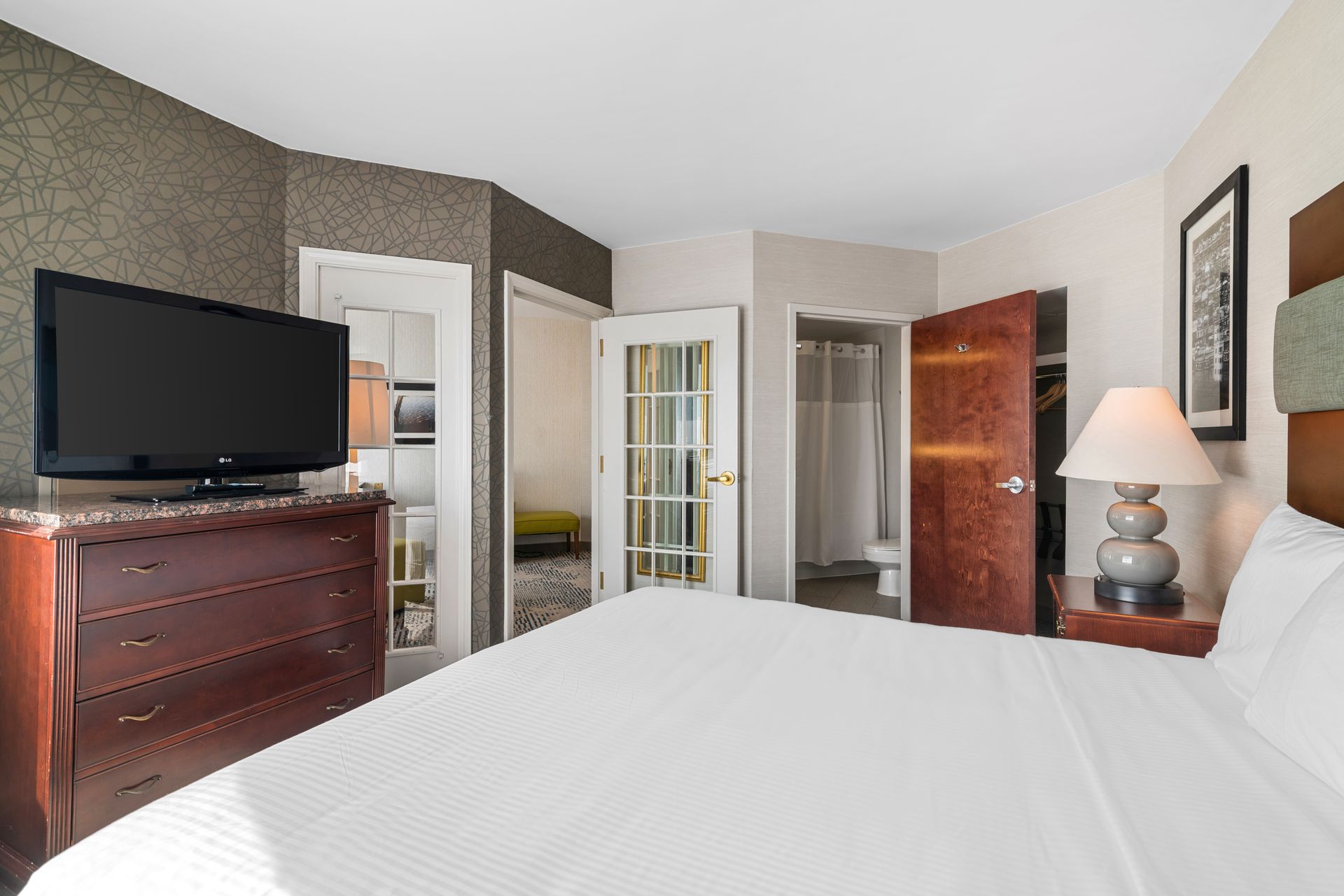 Bright hotel bedroom with white bed, dresser with TV, lamp, open closet, and bathroom door
