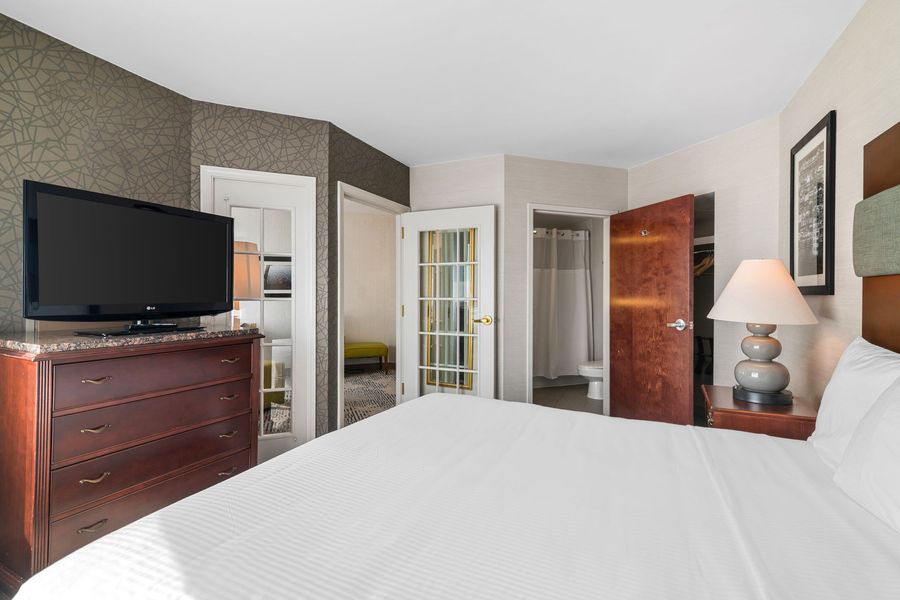Bright hotel bedroom with white bed, dresser with TV, lamp, open closet, and bathroom door