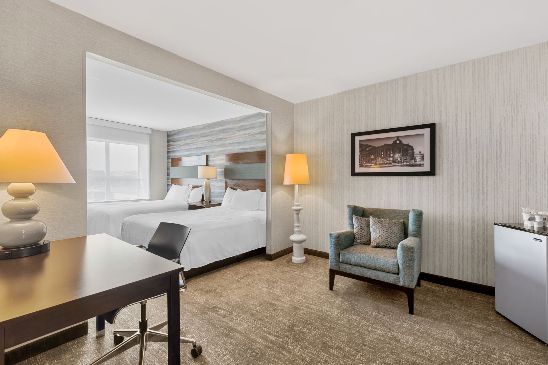 Hotel room with king bed, armchair, desk, lamp, and framed wall art.