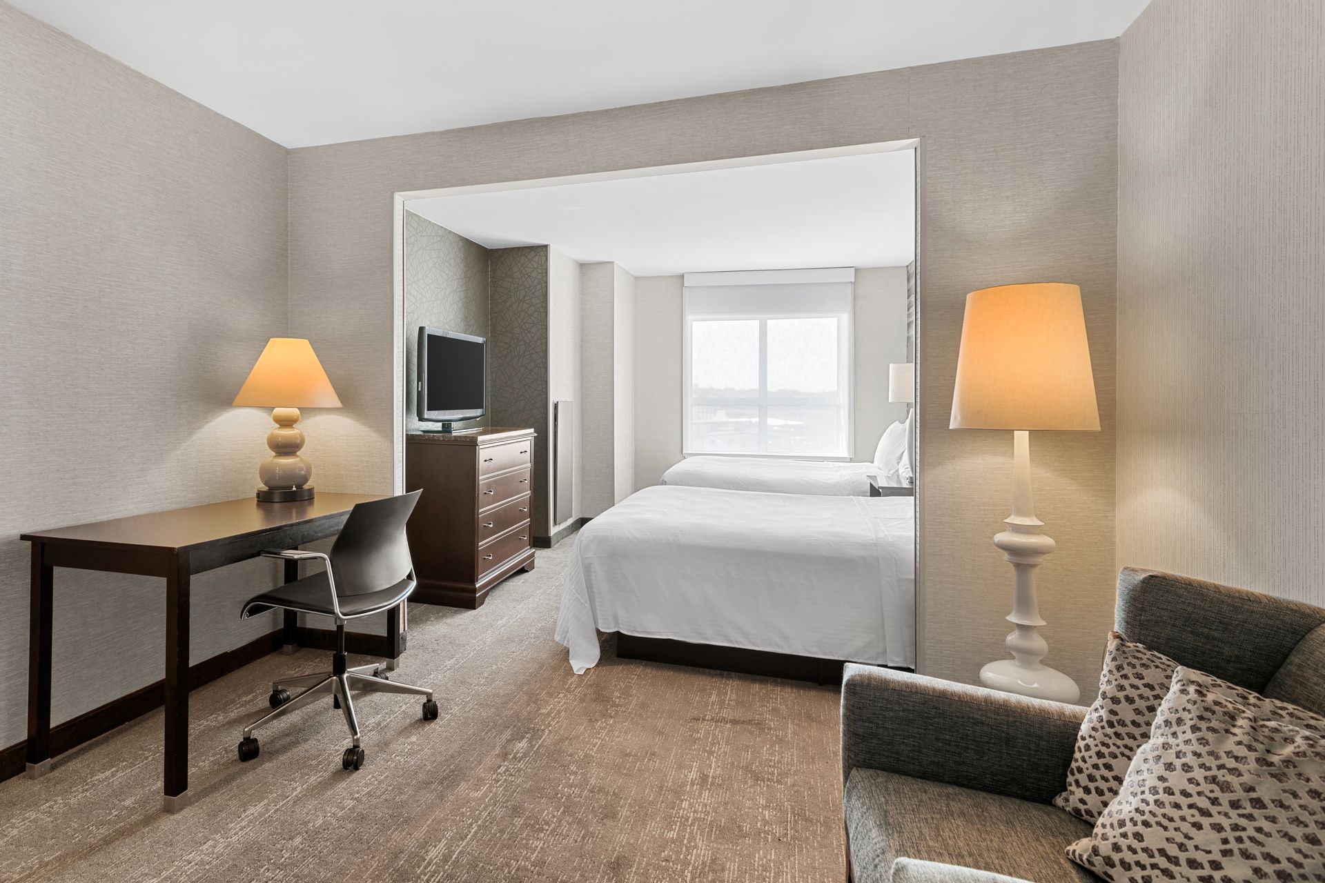 Hotel room with bed, desk, chair, lamps, and seating area in neutral tones