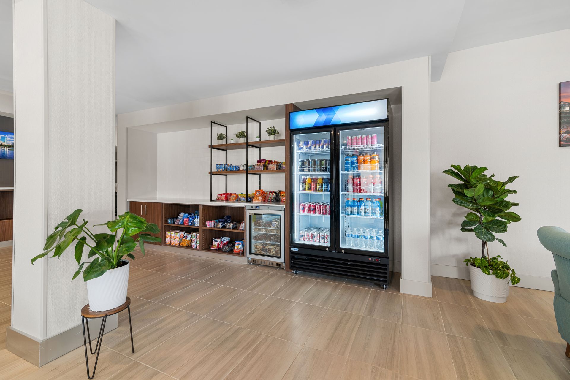 Modern break room with snack shelves, a glass-door fridge, and potted plants on a light wood floor.