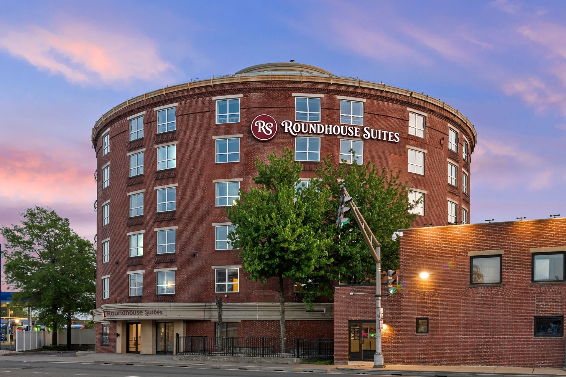 Roundhouse Suites brick hotel at dusk with lit windows and trees in front