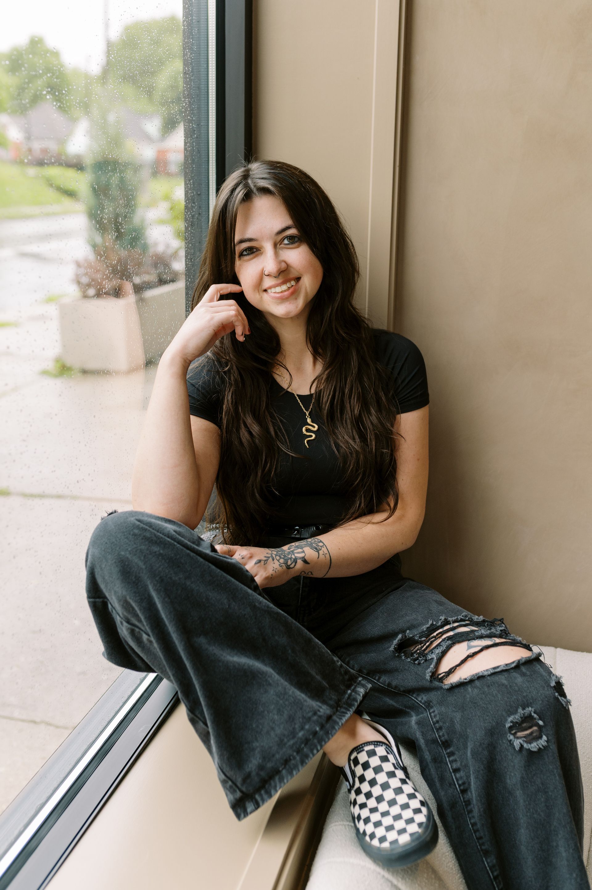 Person sitting by a window in a black top and ripped jeans, smiling with head resting on one hand