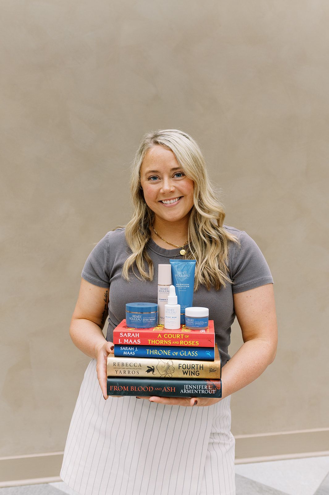 Woman holding a stack of books in front of a beige wall, smiling at the camera.