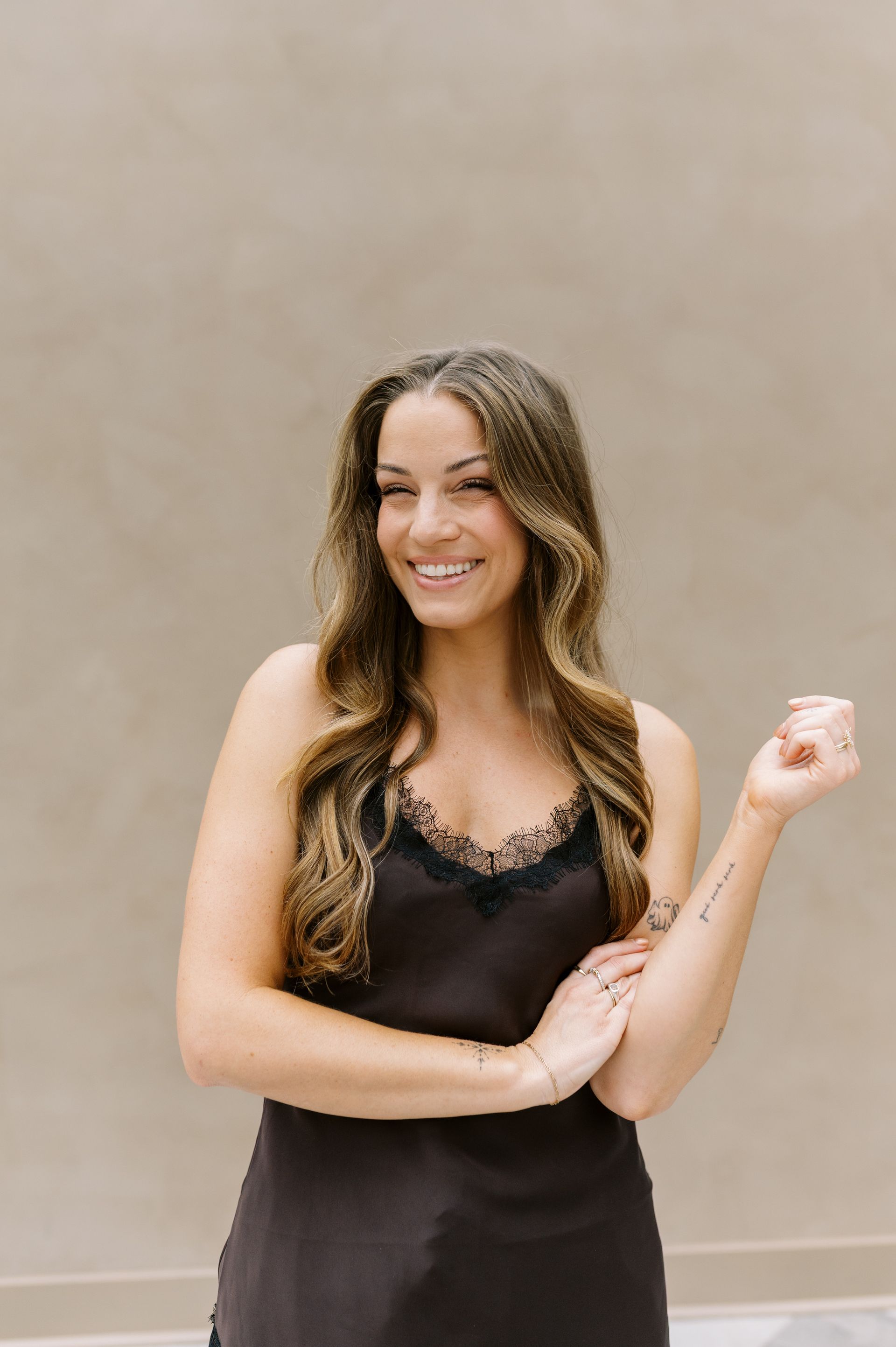 Smiling woman in a black dress posing against a plain beige wall, holding her arm up