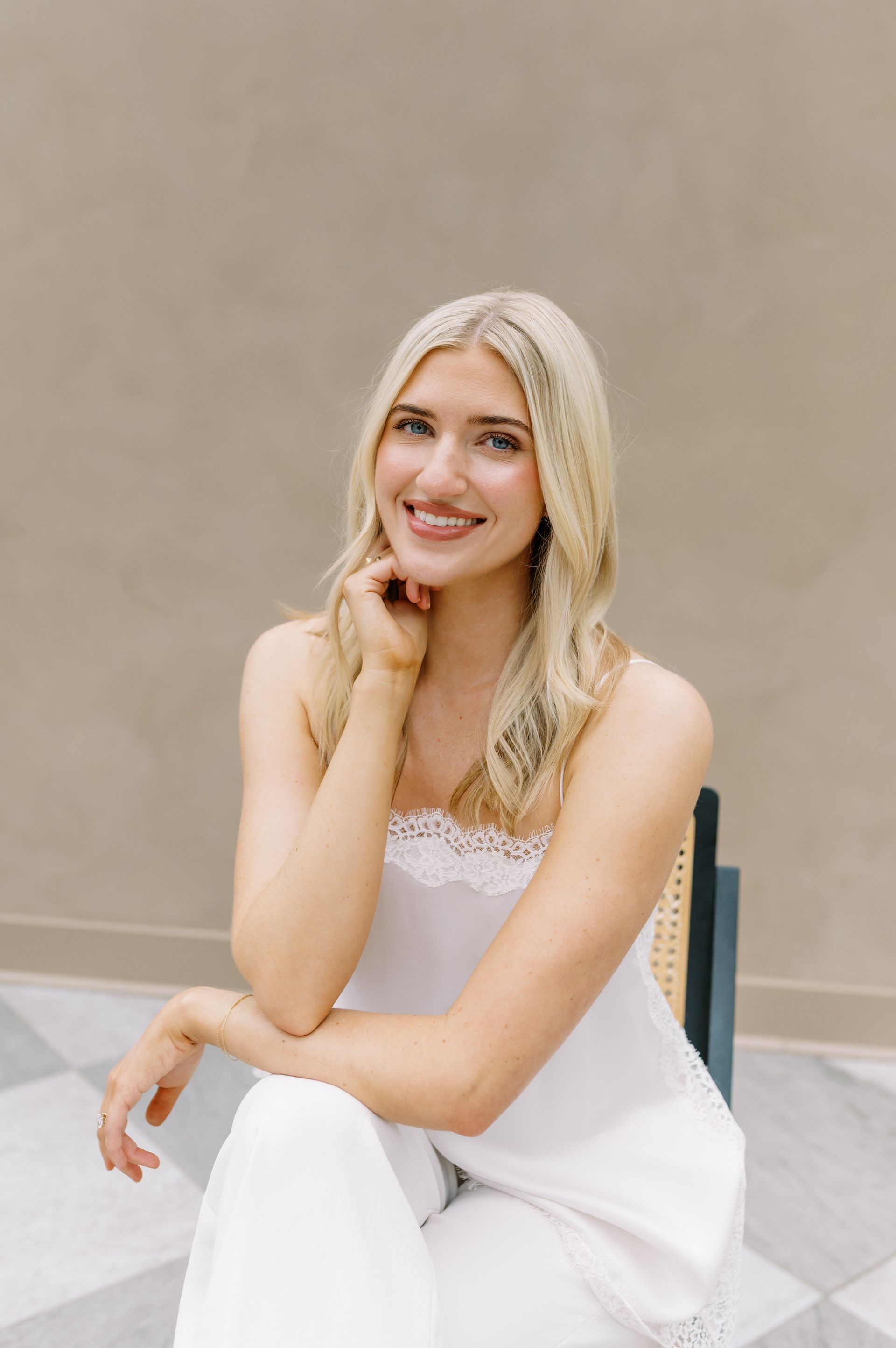 Smiling woman in a white outfit seated against a beige wall, resting her chin on her hand.