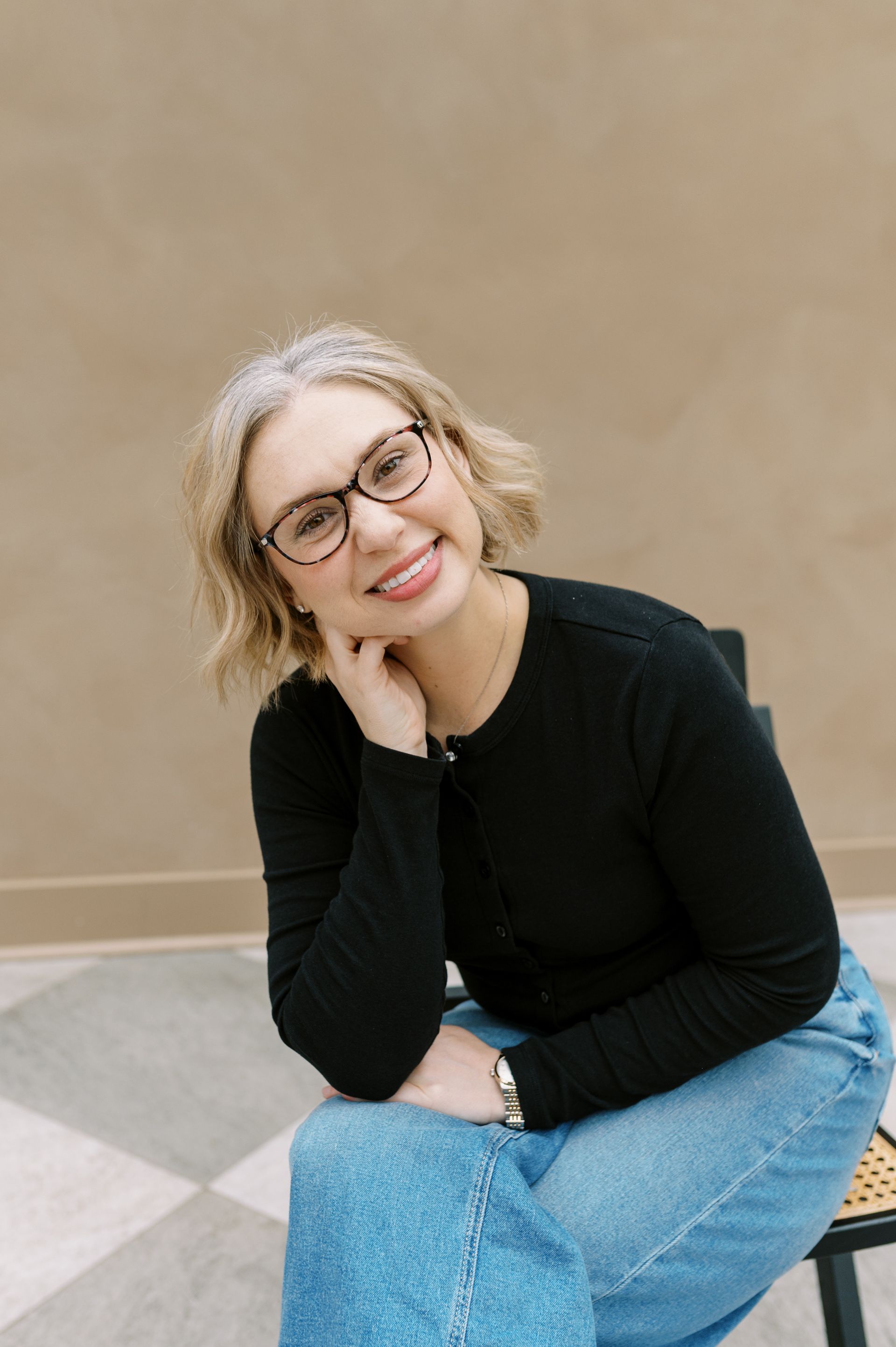 Smiling person in glasses and black top, seated against a beige wall with hand resting on chin.
