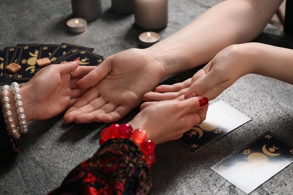 Hands of two people, one examining the other's palm with tarot cards and candles on a table.