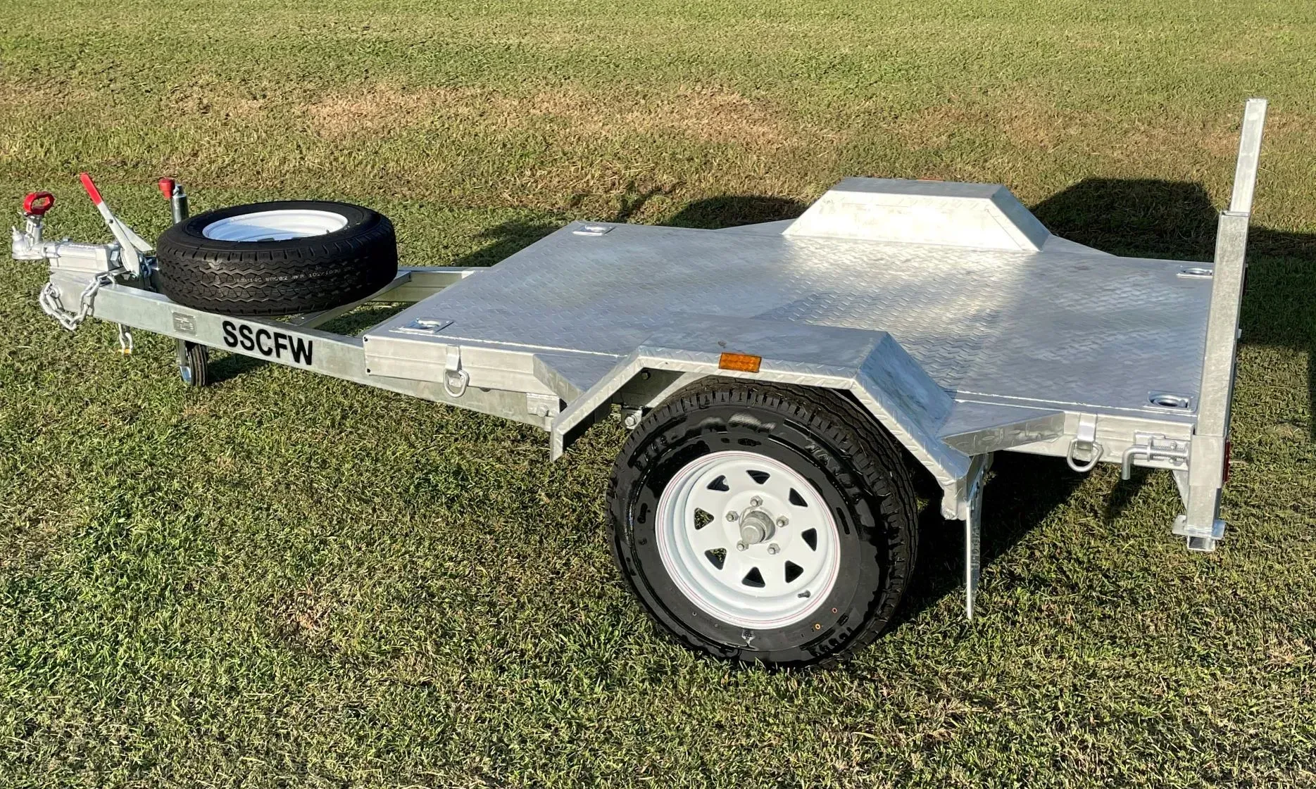 Single-Axle Flatbed Utility Trailer with A Spare Tire, Parked on Grass — SSCFW Trailers In Mareeba, QLD