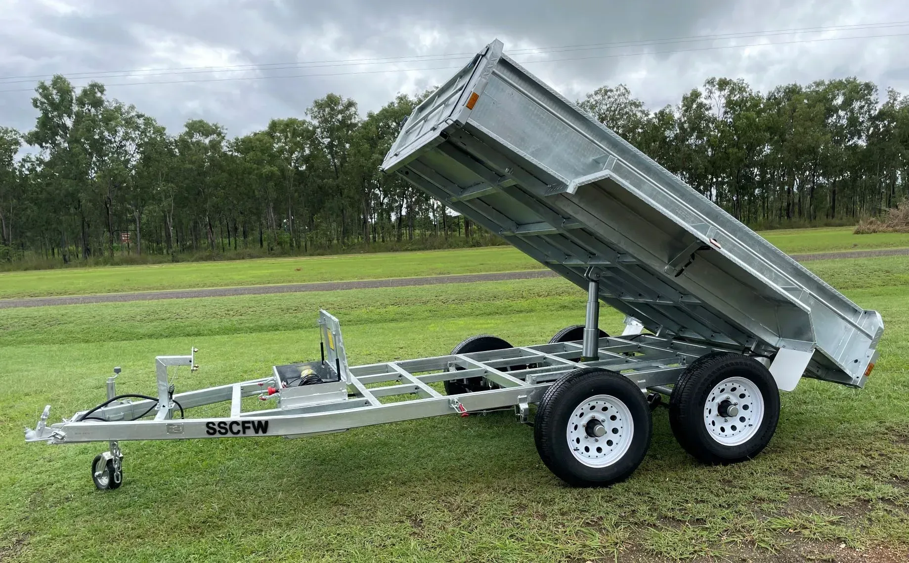Tilt-bed utility trailer with raised rear dump bed on grass under cloudy sky