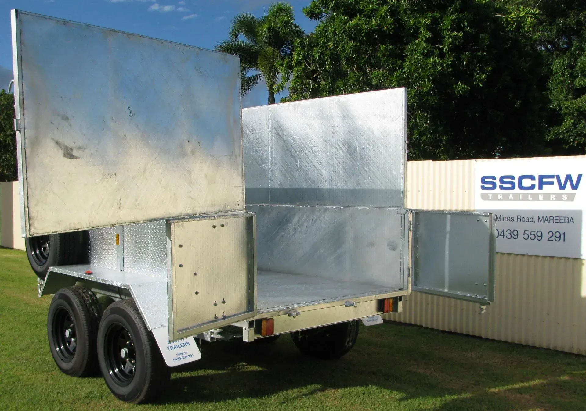 Small Trailer with Large Shiny Metal Panels Parked on Grass Near a Building and Trees — SSCFW Trailers In Cairns, QLD