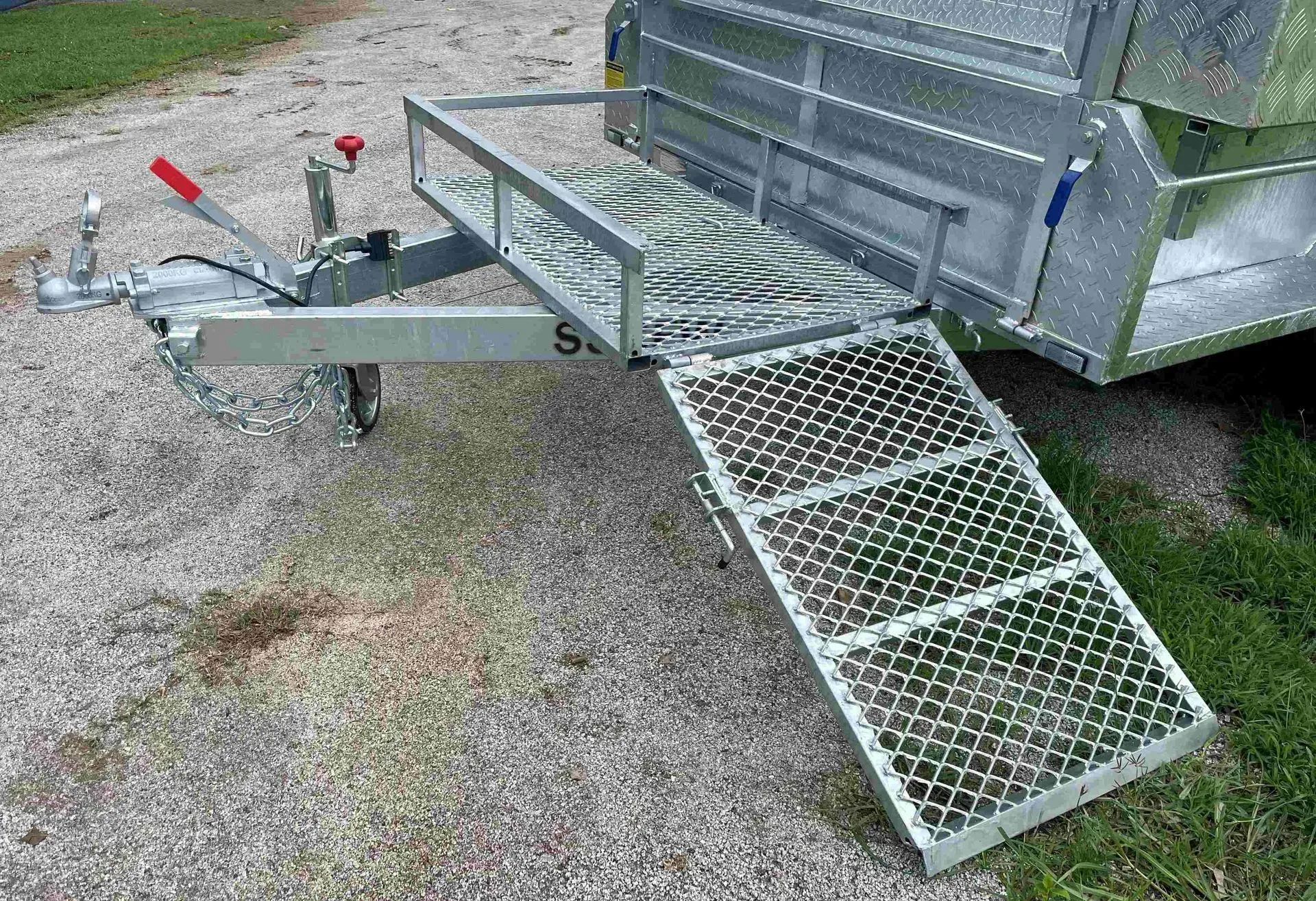 Metal Utility Trailer with A Grated Loading Ramp and Hitch, Parked on Gravel Beside Grass — SSCFW Trailers In Cairns, QLD