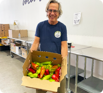 Man smiling, holding a box of colorful peppers. He's inside a brightly lit warehouse.