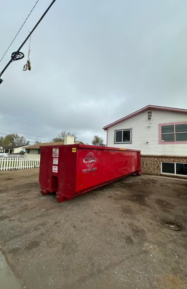 Red dumpster in a driveway next to a white house with pink trim and a cloudy sky overhead.