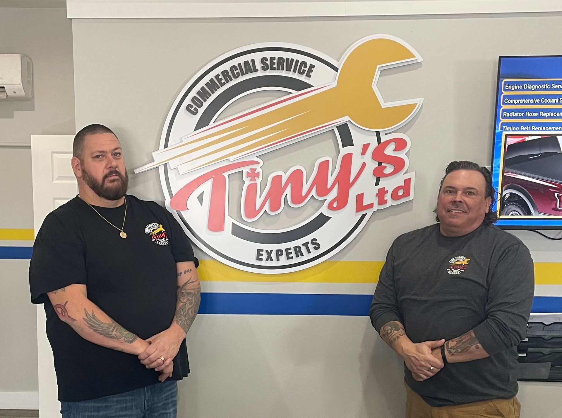 Two men are standing in front of a sign for tiny 's ltd. | Tiny's Automotive