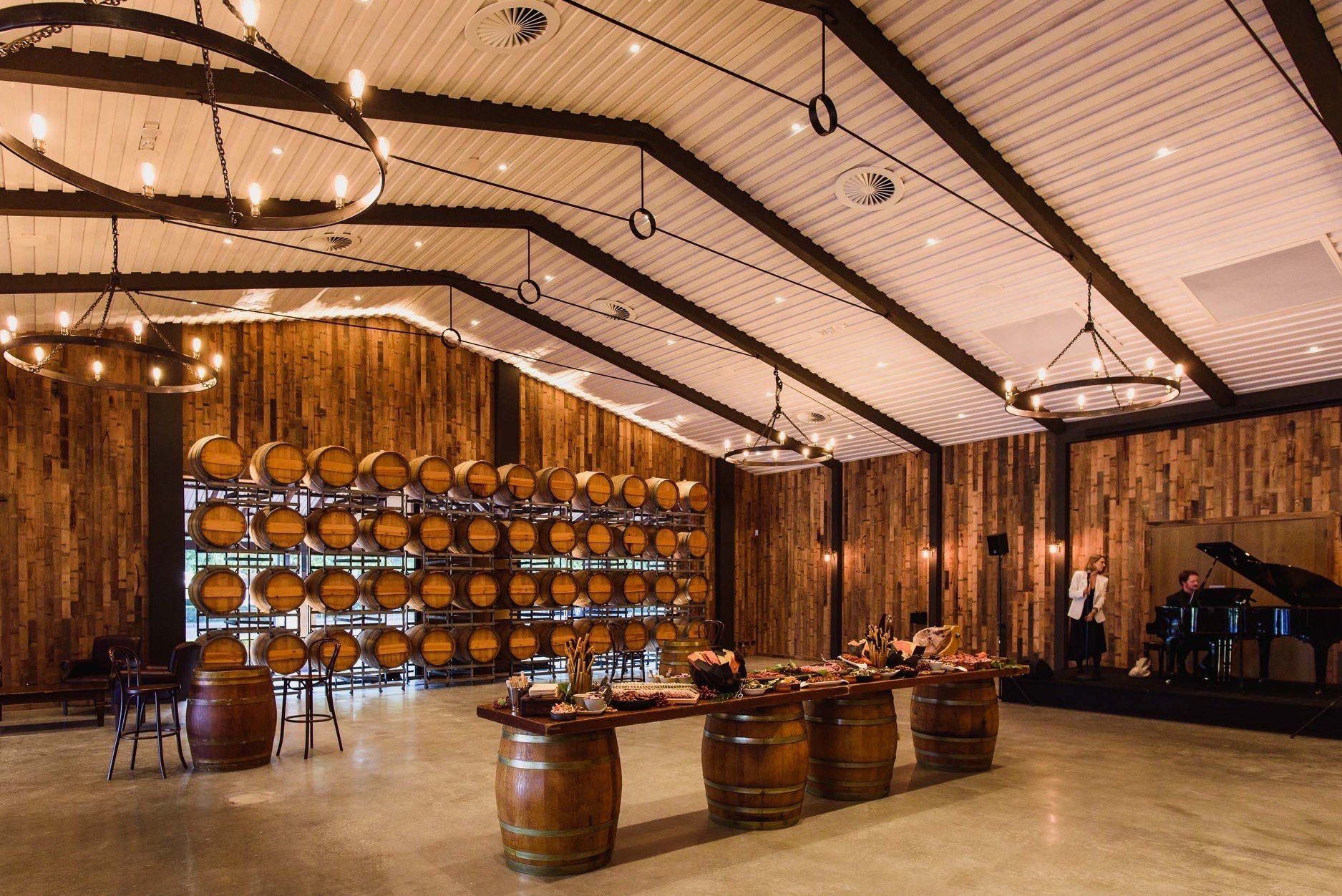 Winery event space with timber walls, stacked wine barrels, barrel tables, banquet food display and a stage with a grand piano.