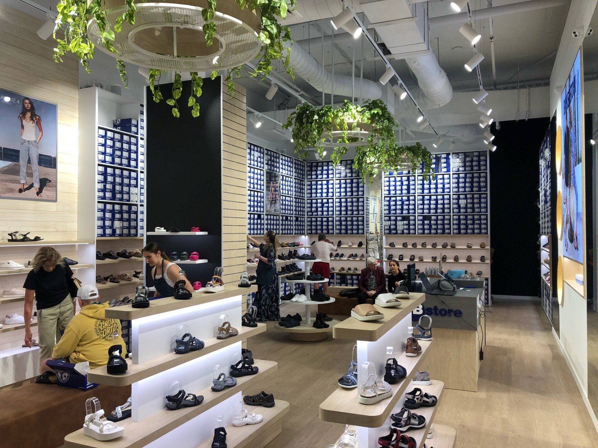 Birkenstock footwear store with illuminated shoe displays, hanging greenery, wall shelving and customers browsing sandals and shoes.
