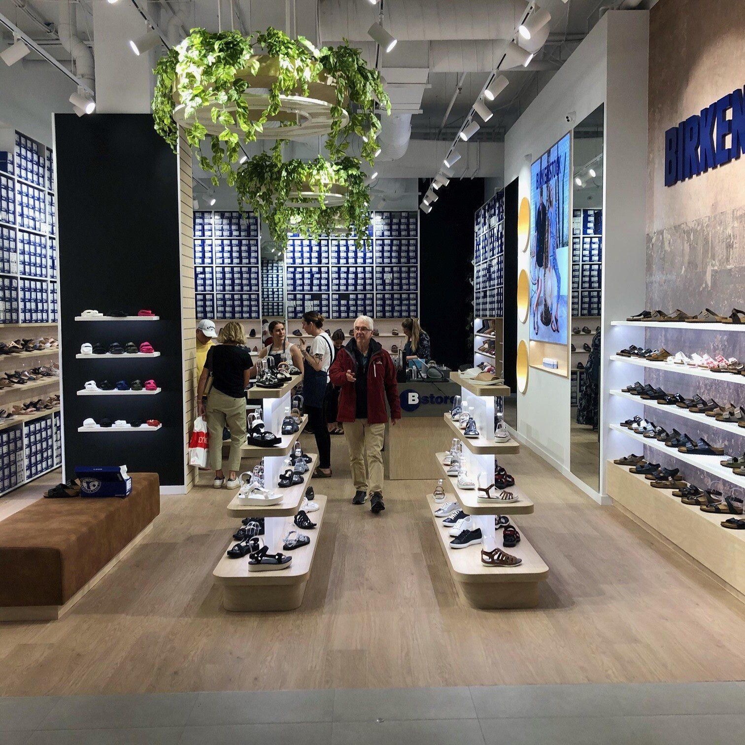 Birkenstock footwear store interior with sandal displays, wall shelving, hanging plants and customers browsing products.
