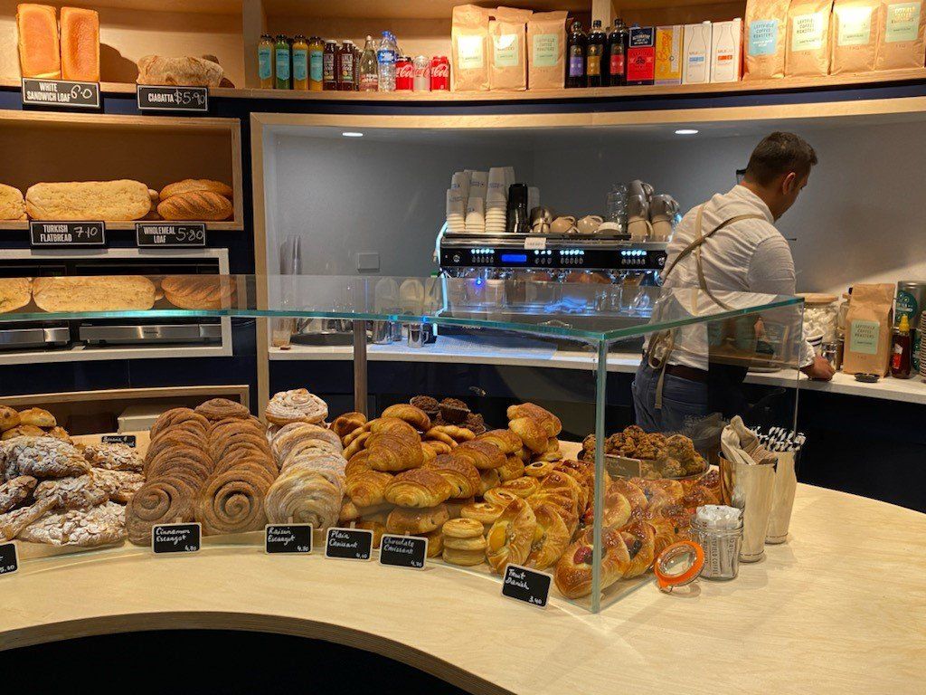 Bakery display counter with assorted pastries, croissants and baked goods, while a staff member prepares coffee behind the counter.