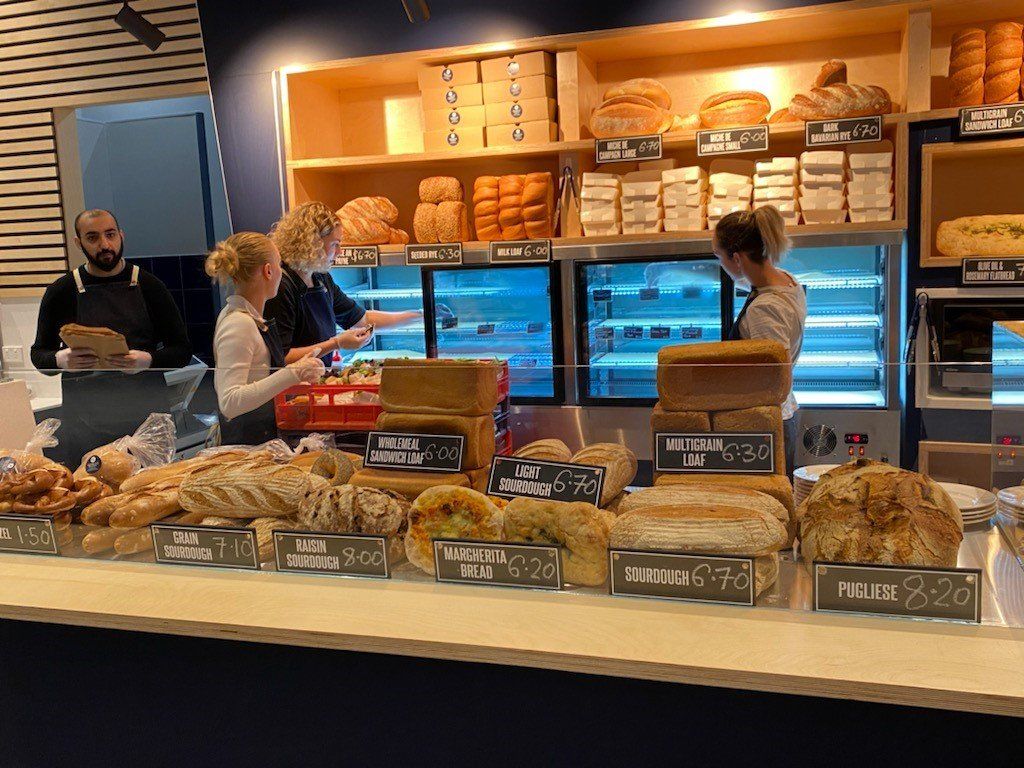 Bakery counter displaying artisan bread loaves and baked goods with staff serving customers behind the display cabinet.