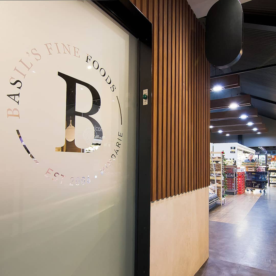 Entrance area of Basil’s Fine Foods featuring branded glass signage, timber wall panelling and access to the supermarket interior.