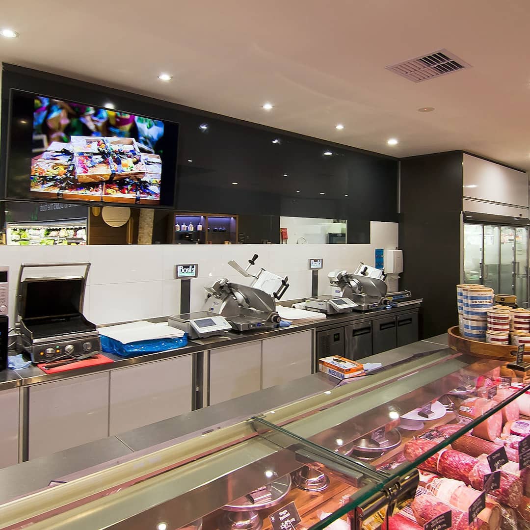 Deli counter with cured meats, food preparation equipment and refrigerated displays inside a modern gourmet food store.