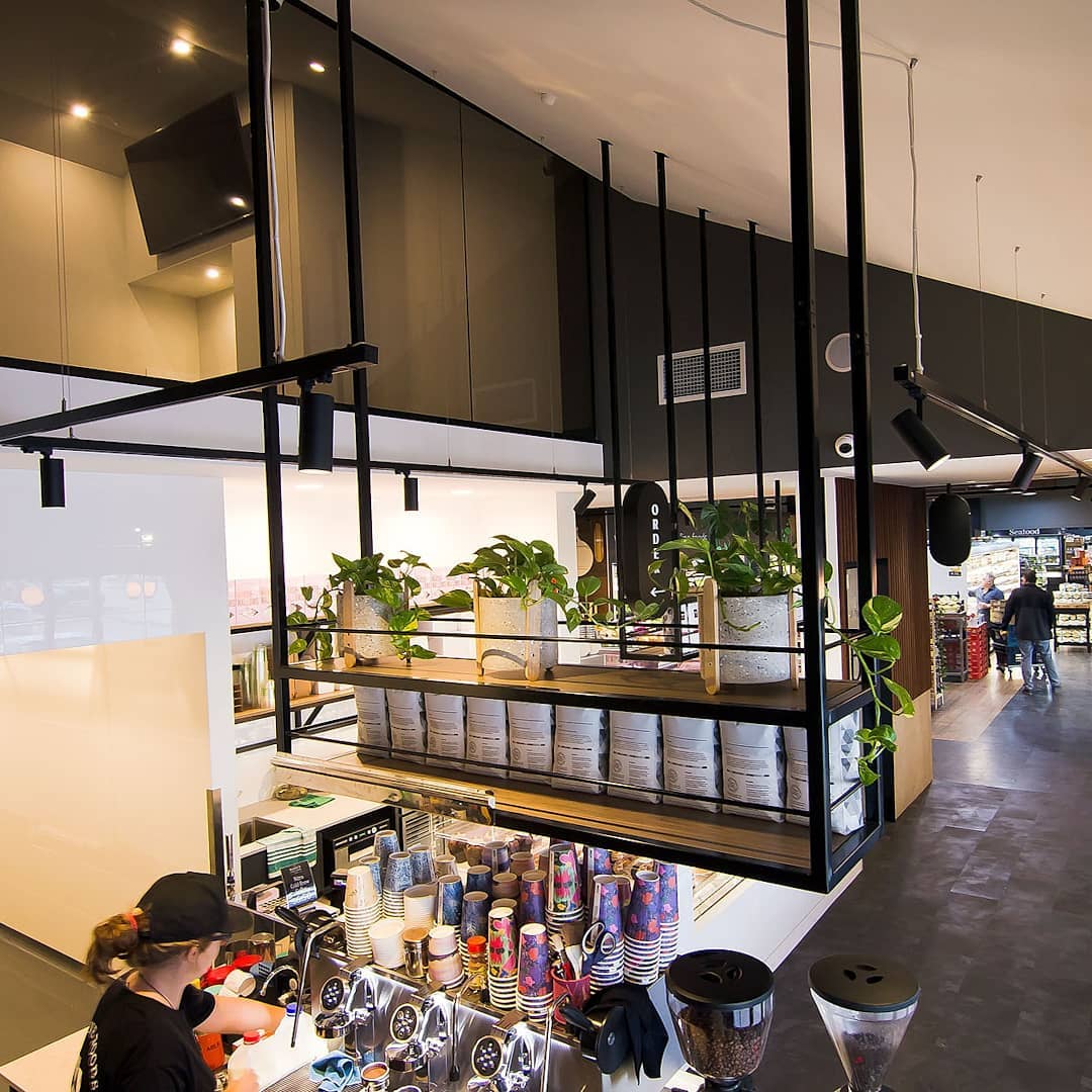 Coffee preparation area with espresso machines, takeaway cups, hanging shelves and indoor plants inside a modern café.
