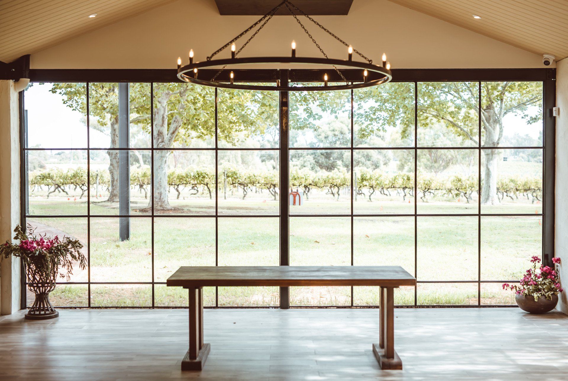 Event venue interior with a timber table, large floor-to-ceiling windows overlooking vineyards and a circular chandelier hanging from the ceiling.
