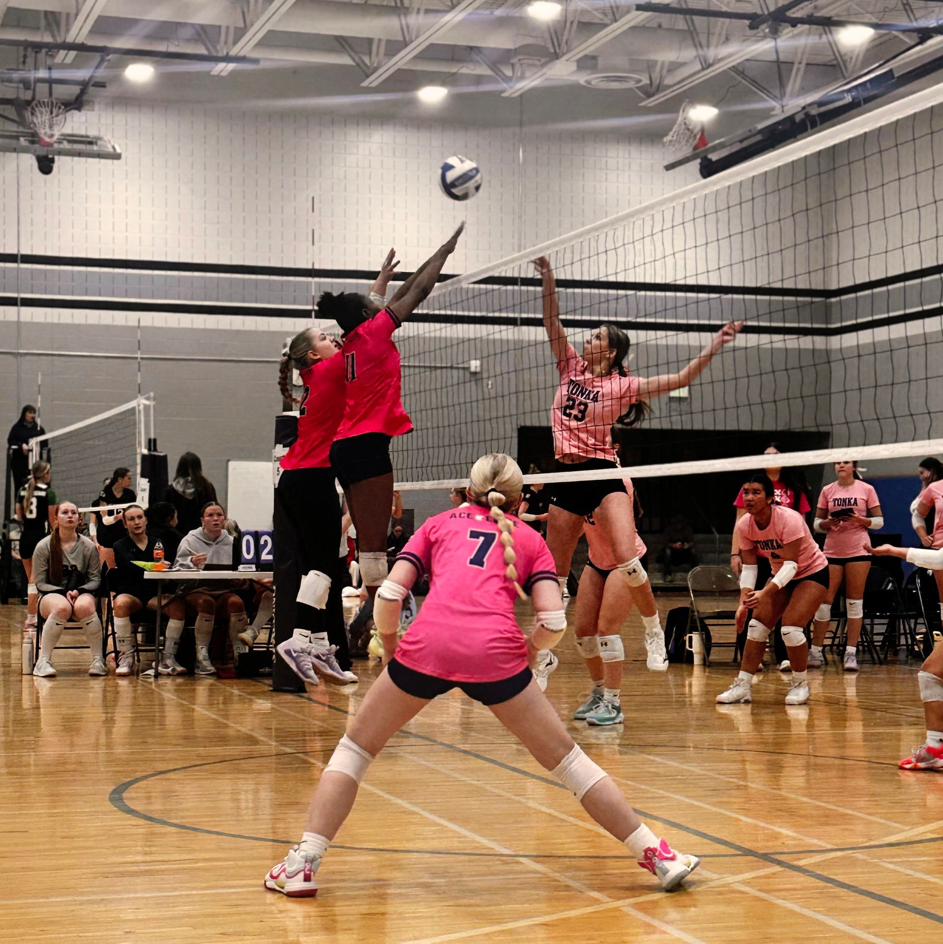 Indoor volleyball game with players in pink and red jumping near the net as the ball drops over the court