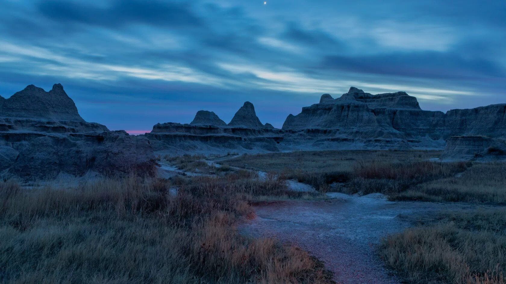 Badlands landscape at dusk: rugged rock formations, blue sky, path, and tall grass.