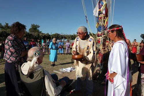 Native American ceremony with elders and attendees outdoors, near a decorated pole.